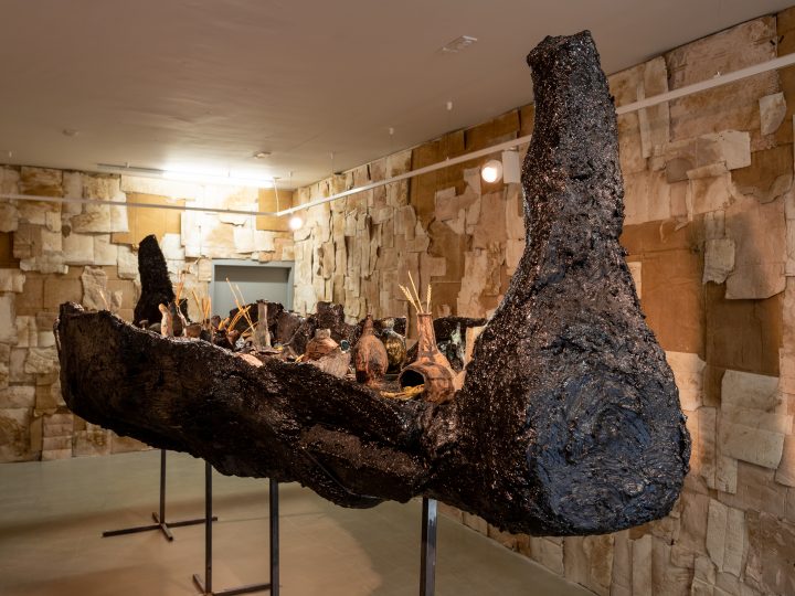 Image of artist Emii Alrai's show 'The High Dam. A 5ft boat made of bitchumen stands in a room with small clay pots inside the boat. The walls of the room are covered in squares of cardboard.