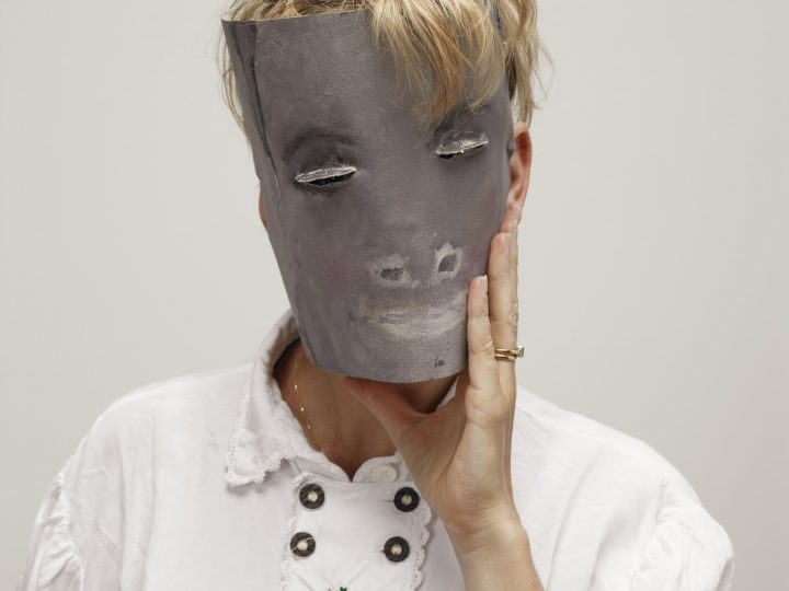 Artist Laure Prouvost is photographed with a paper mask covering her face. The mask is gray with painted features including nose, mouth and eyes which are small slits. She wears a white shirt and has blonde hair.