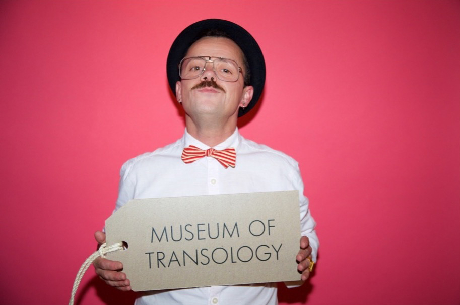 In this image, E-J is a masculine presenting trans man with a moustache and wears gold spectacles, a black trilby hat, a cream and pink striped bowtie and pink business shirt. He is solding up a large brown Museum of Transology sign that is an oversized version of the tags that are attached to the objects in the MoT collection.