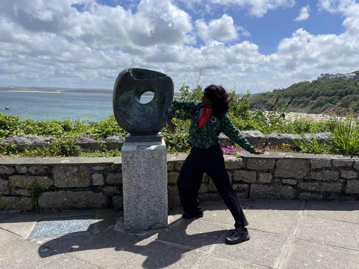 A woman (Nwando) poses next to a Barbara Hepworth sculpture on a sunny day near the sea.